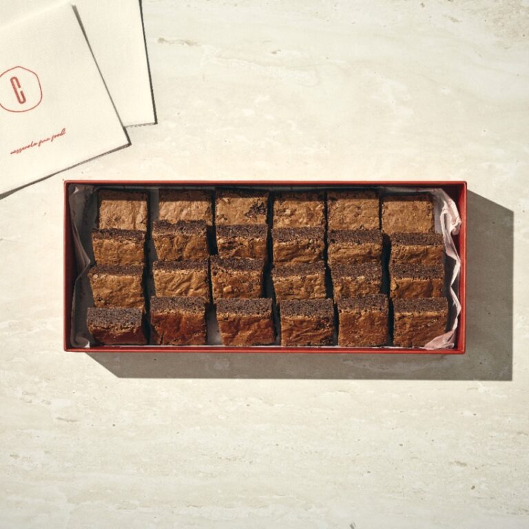 קופסה מלבנית של בראוניז, המכילה שש עשרה חתיכות בראוניז חתוכות באופן אחיד, מונחת על משטח אבן בהיר לצד מספר מפיות מקופלות. - Cassarole