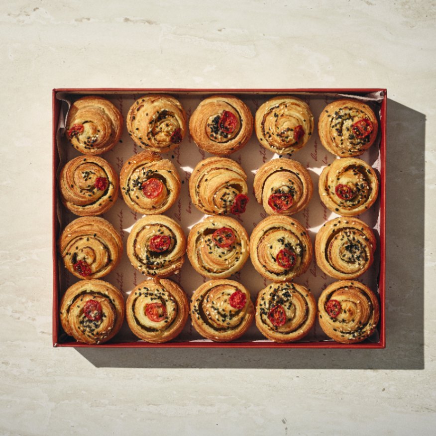 קופסה של ברושים מלוחים: עשרים לחמניות מלוחות אפויות, חלקן עם חצאי עגבניות שרי ואחרות עם זרעי שומשום שחורים, מסודרות בקפידה על נייר אפייה. - Cassarole