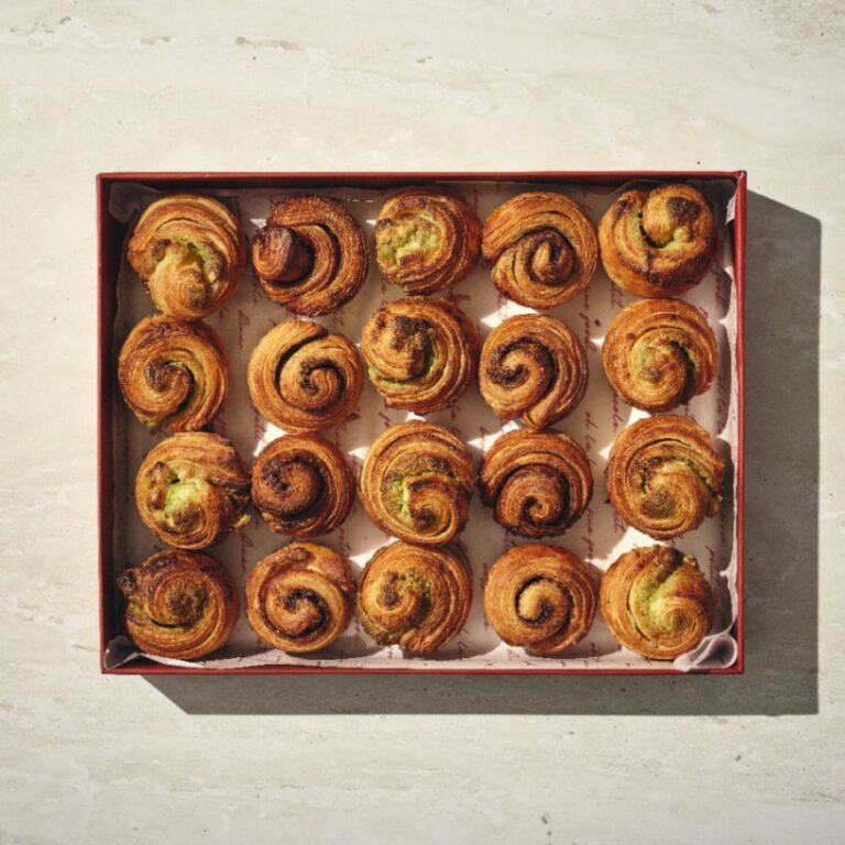 קופסה של בריושים מתוקים — שש עשרה מאפים בצבע חום-זהוב, בצורת ספירלה — מסודרים ברשת 4x4 על נייר אפייה. - Cassarole