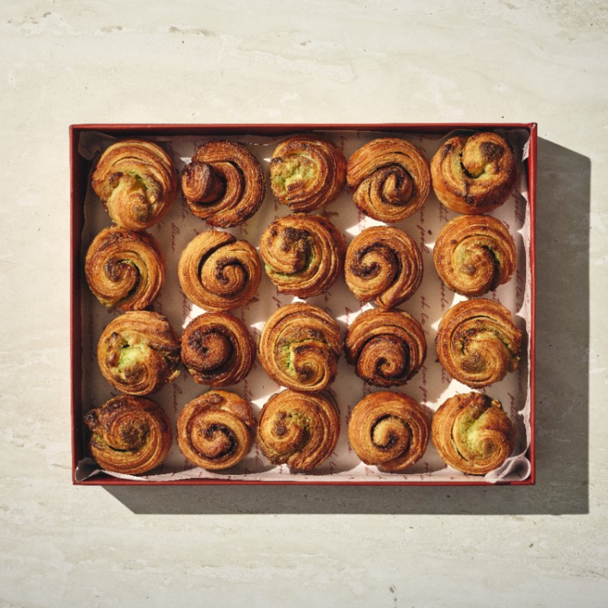 קופסה של בריושים מתוקים — שש עשרה מאפים בצבע חום-זהוב, בצורת ספירלה — מסודרים ברשת 4x4 על נייר אפייה. - Cassarole