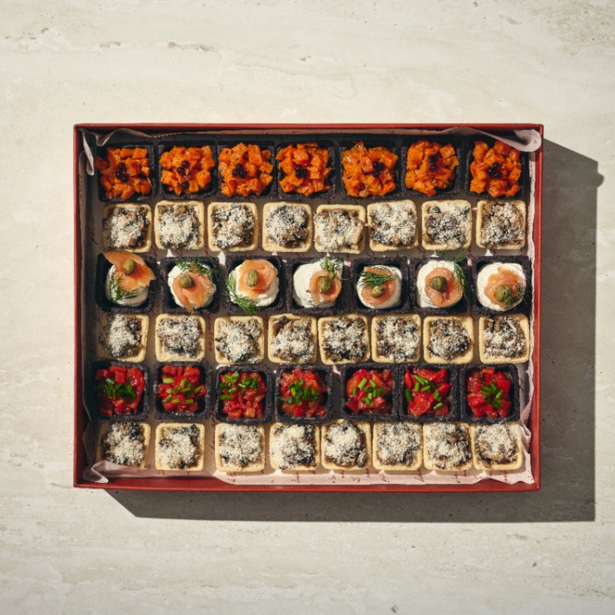 קופסה של טארטלט שחור המכילה מגוון מתאבנים בגודל ביס, המסודרים בקפידה ברשת ומצופים בעשבי תיבול, ירקות וממרחים. - Cassarole