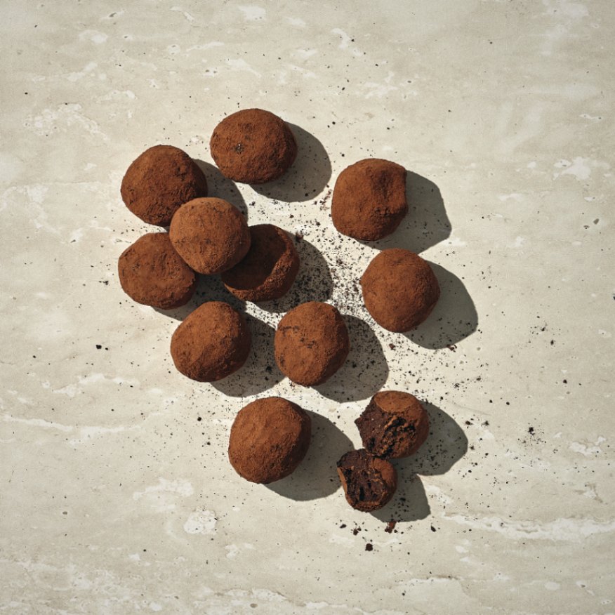 קבוצת טראפלס שוקולד מצופים באבקת קקאו מסודרים על משטח בהיר ומחוספס, עם כמה פירורים מפוזרים סביבם. - Cassarole