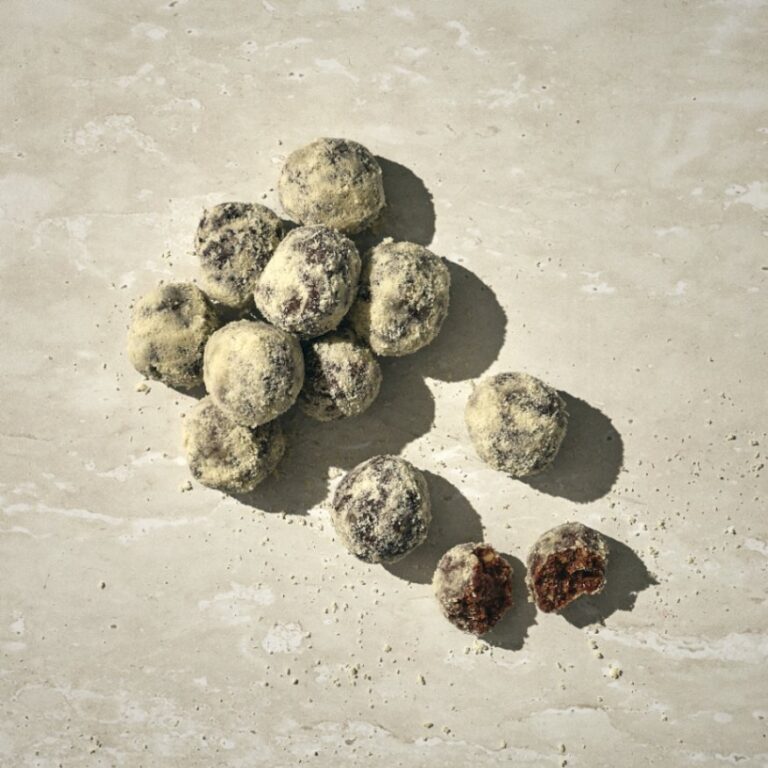 קבוצת כדורי תמרים מסודרים על משטח אבן בהיר, זרועים אבקת סוכר, כאשר אחד מהם חתוך וחושף את המילוי הכהה שלו. - Cassarole