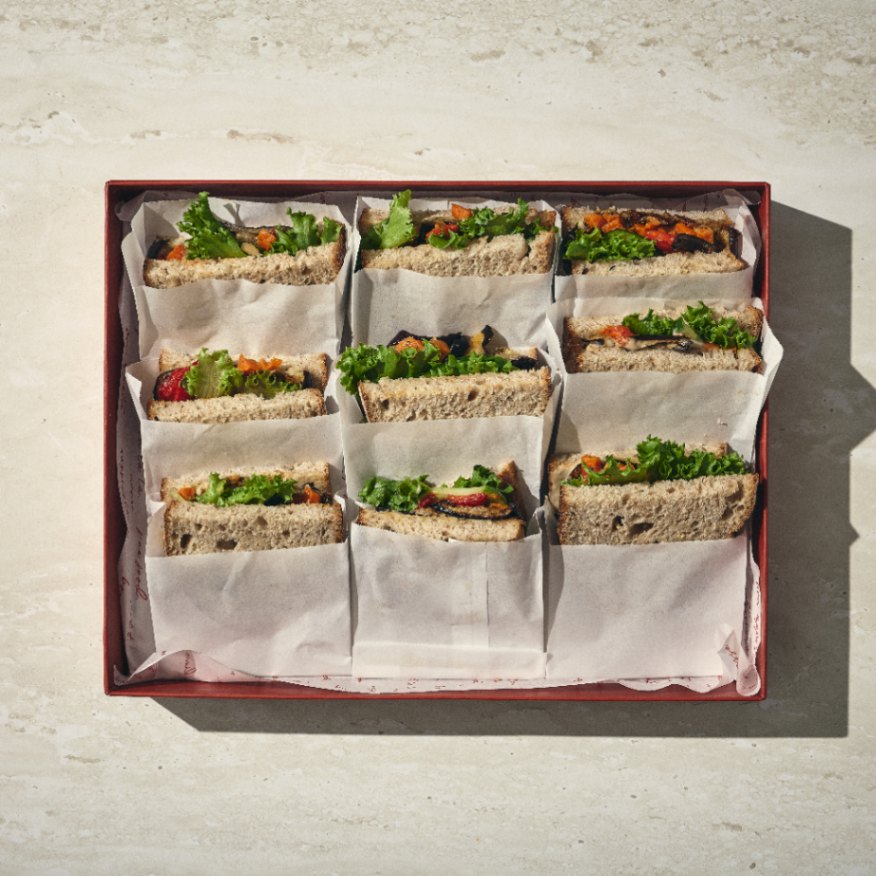 קופסה של תשעה כריכים טבעוניים, כל אחד מהם מלא בעלים ירוקים וירקות נראים לעין, מסודרים בקפידה ומופרדים בנייר אפייה. - Cassarole