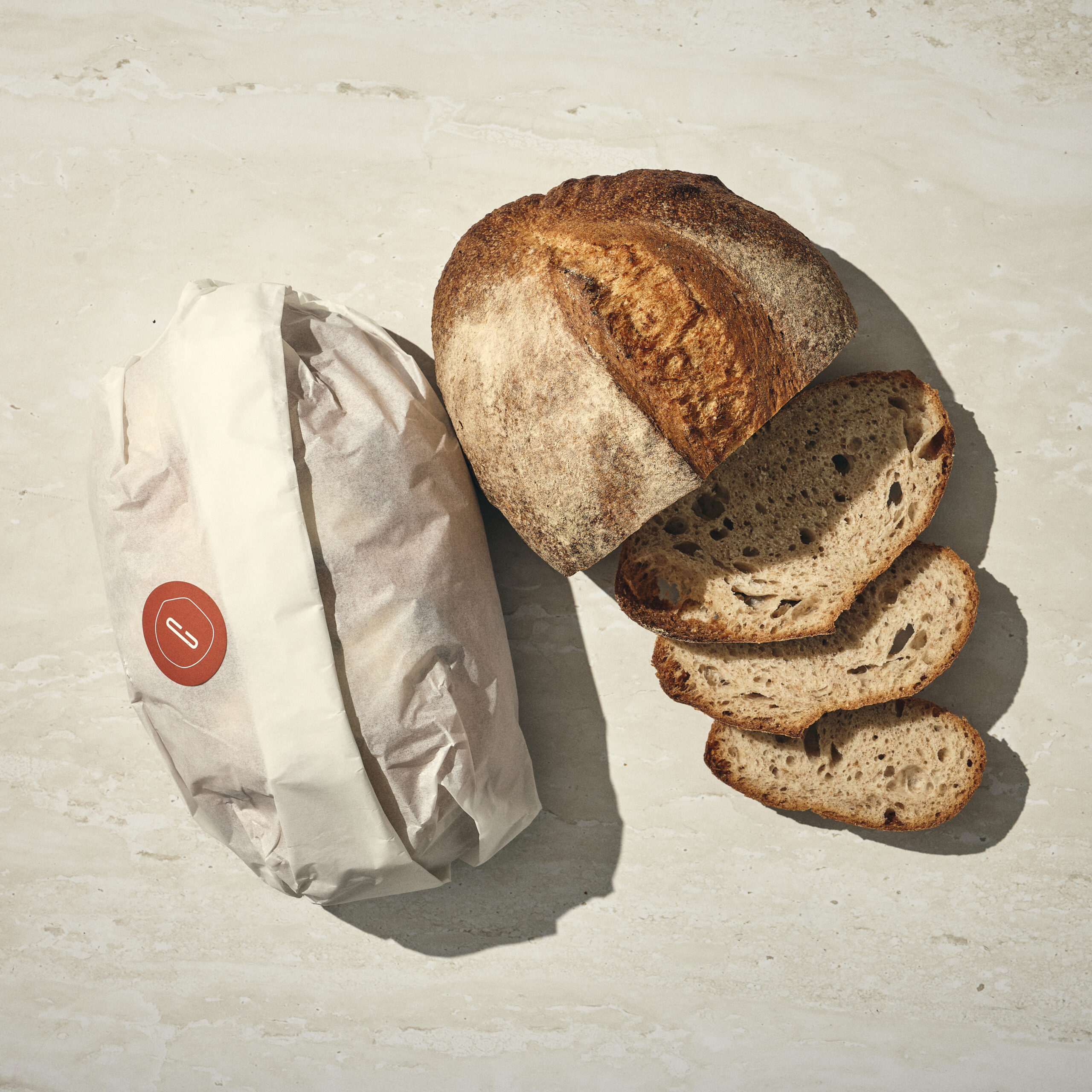 כיכר לחם מחמצת חתוכה חלקית מונחת לצד פריט עטוף על משטח בהיר. - Cassarole