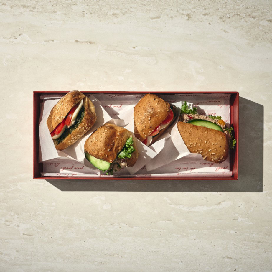 ארבעה כריכים קטנים עם לחמניה ללא גלוטן, חסה, עגבנייה ומלפפון מסודרים במגש מלבני על משטח בהיר. - Cassarole