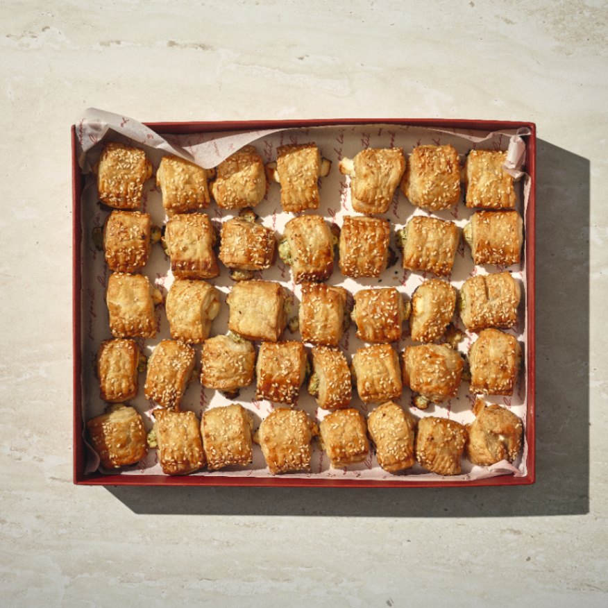 קופסה של מאפים קטנים מלוחים: חמש שורות של מאפים מרובעים בצבע חום-זהוב, כל אחד מהם מצופה בשומשום ומסודר בקפידה על נייר אפייה. - Cassarole