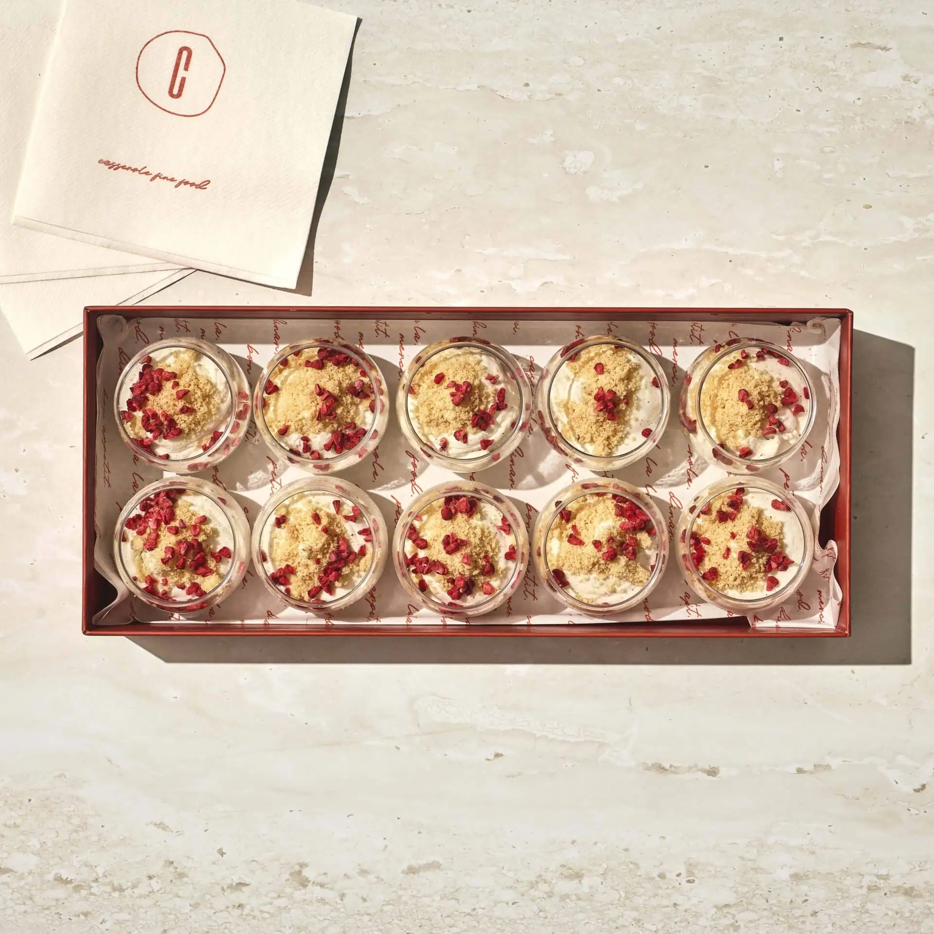 קופסה של עשרה גביעי מוס מסקרפונה עם פירורים מעל מונחת על משטח בהיר לצד מפיות ממותגות. - Cassarole