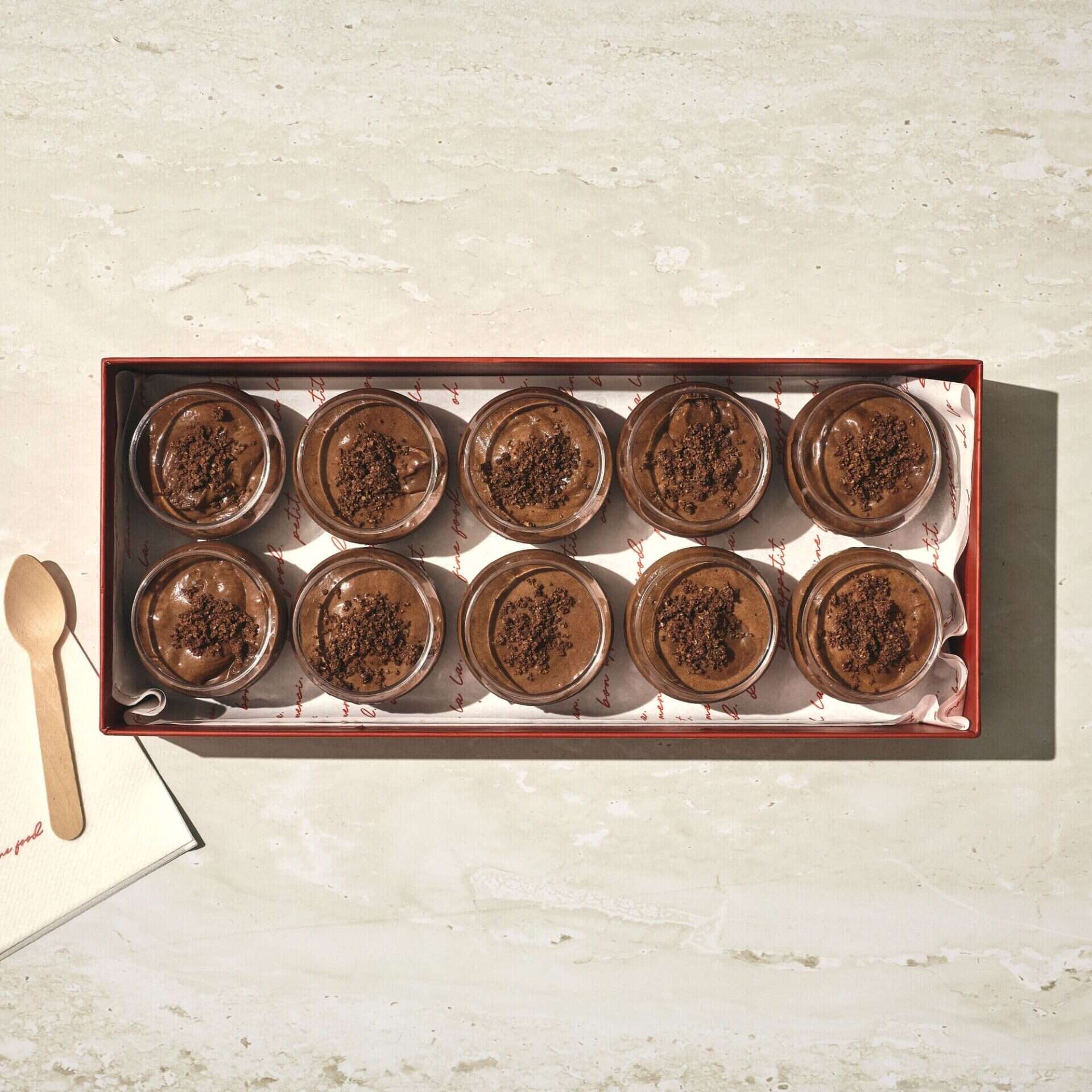 מגש מלבני מציג עשרה כוסות זכוכית של מוס שוקולד, כל אחת מהן מעוטרת בשבבי שוקולד. כף עץ ומפית מונחות משמאל למגש. - Cassarole