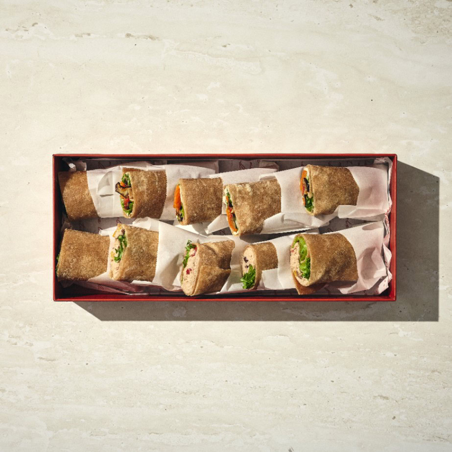 קופסה מלבנית מכילה עשרה מיני טורטיות טבעוניות, לחמניות סנדוויץ' עטופות עם ירקות ירוקים ומילויים אחרים, מסודרות בקפידה על נייר אפייה. - Cassarole