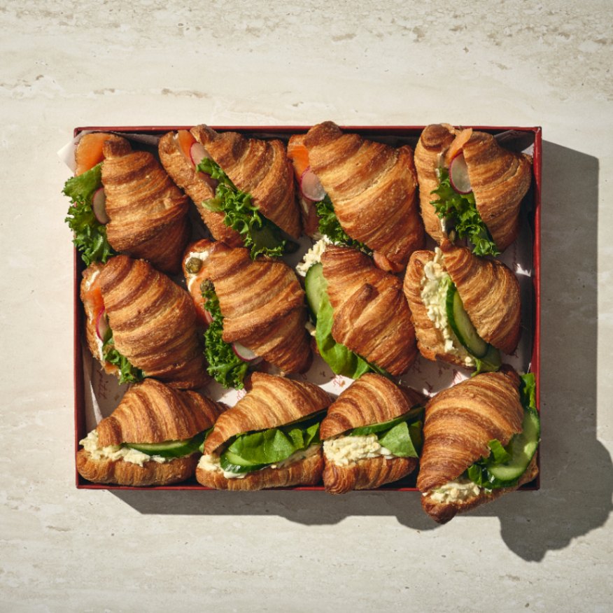 קופסה של מיני קרואסון — תשעה קרואסונים ממולאים בחסה, עגבנייה ומילויים שונים — מסודרים בשלושה טורים על משטח בהיר. - Cassarole