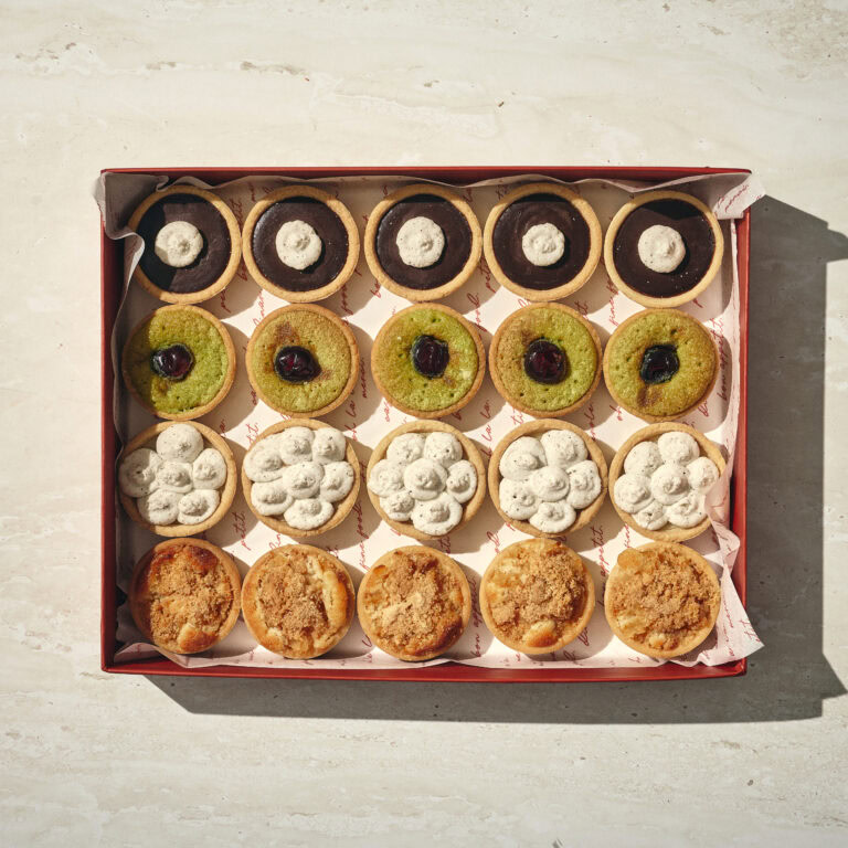 קופסה של מיקס טארטלטים: שש עשרה טארטלטים קטנים ומגוונים בארבע שורות, כל אחד עם ציפוי ייחודי, מסודרים על נייר אפייה. - Cassarole