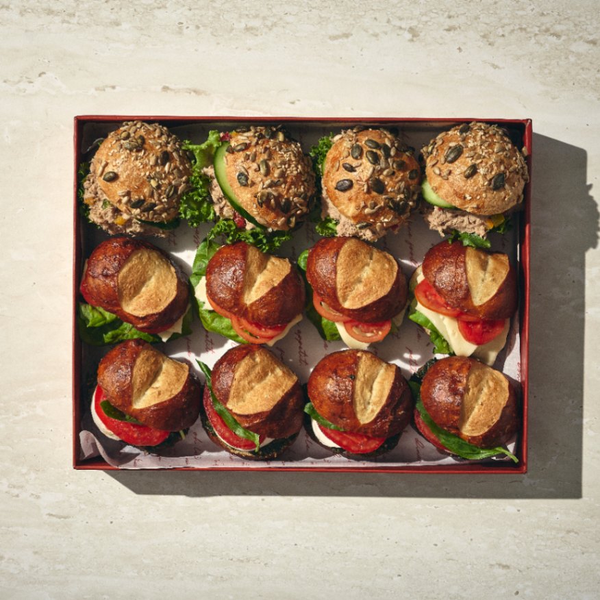 מיקס כריכים: קופסה עם שישה מיני סנדוויצ'ים בלחמניות שומשום בשורה העליונה ושישה מיני סנדוויצ'ים בלחמניות פרצל בשורה התחתונה, כולם עם חסה ומילויים גלויים. - Cassarole