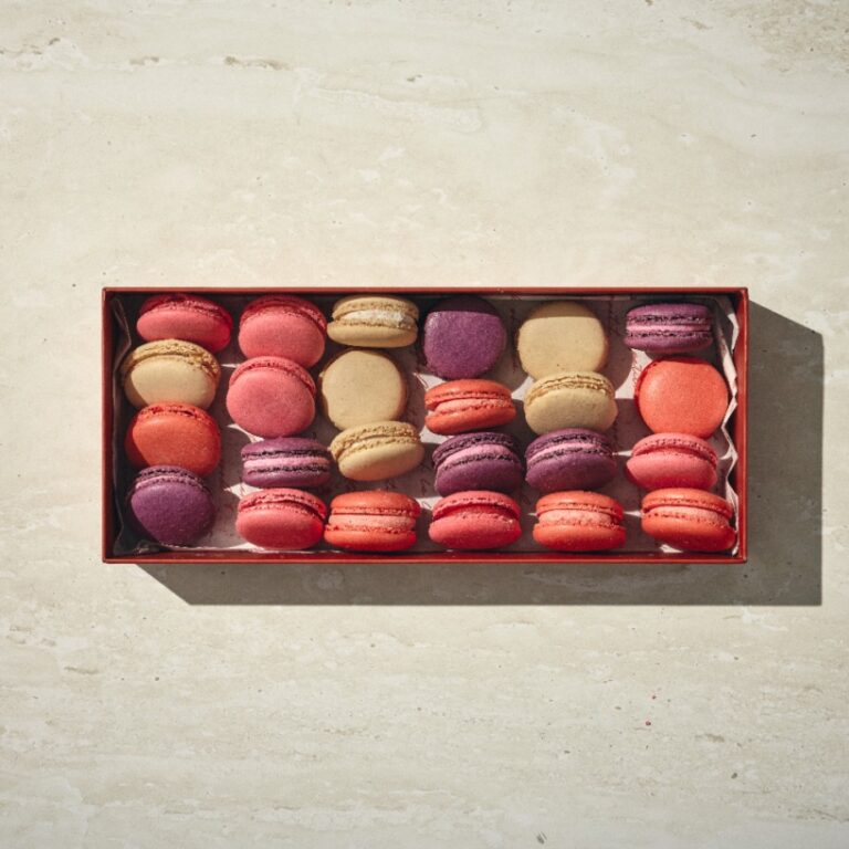 קופסה מלבנית של מקרון צרפתי בצבעים אדום, סגול ובז' מוצגת על משטח אבן בהיר. - Cassarole