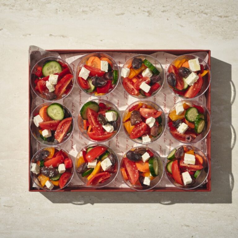 מגש עם 12 כוסות סלט יווני אישי, כל אחת ממולאת בסלט יווני המורכב מעגבניות, מלפפונים, זיתים וגבינת פטה על משטח בהיר. - Cassarole