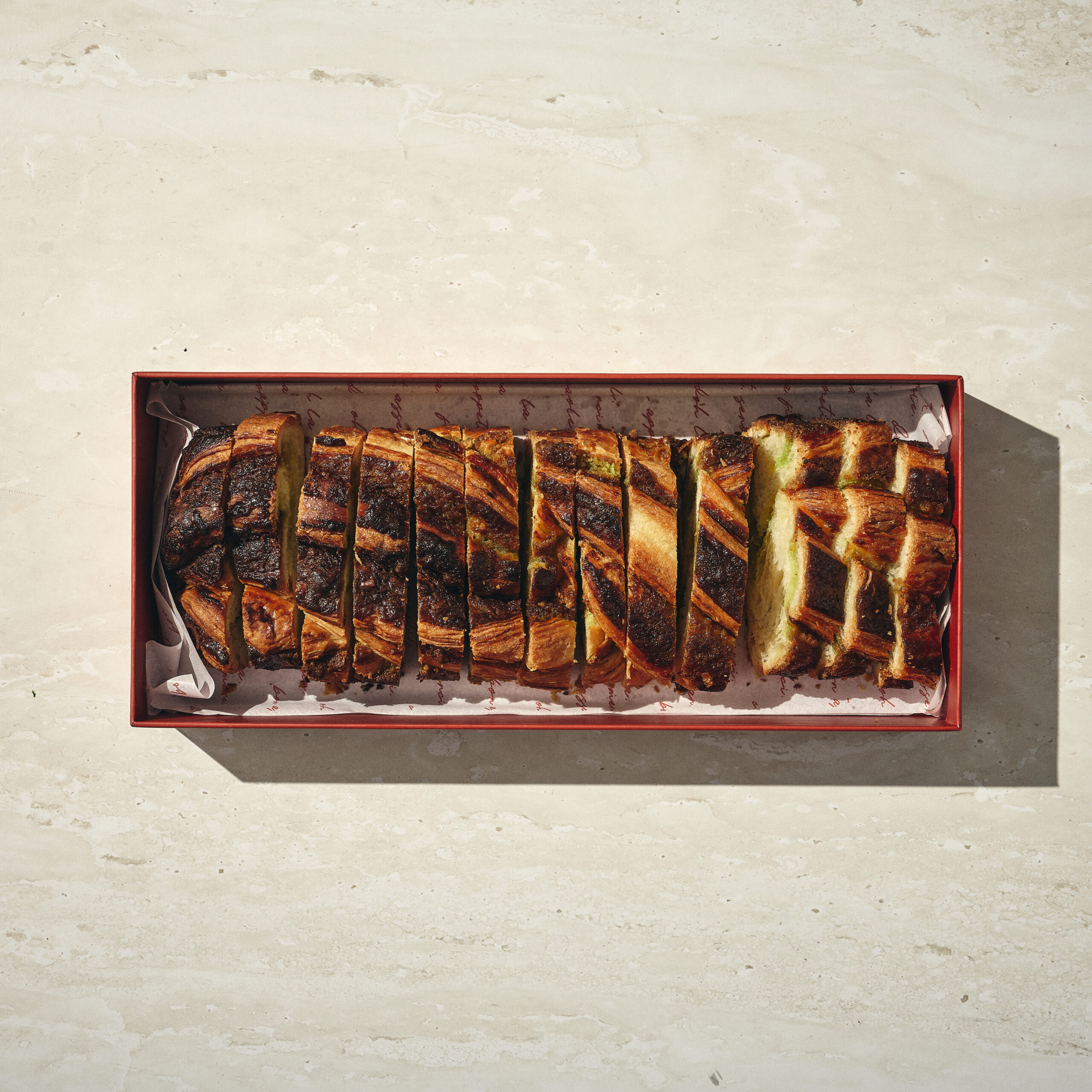 קופסה מלבנית עם עוגת קראנץ' פיסטוק, בעלת קרום חום-זהוב ומרופדת בנייר אפייה, מונחת על משטח בהיר. - Cassarole