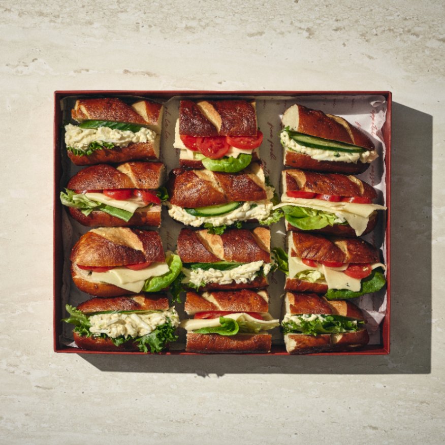 קופסה בגודל 3x3 של פרעצלס — תשעה חצאי כריכים עם לחמניות פרעצל, חסה, עגבנייה, גבינה ומילוי שמנת — מסודרים בקפידה על משטח בהיר. - Cassarole