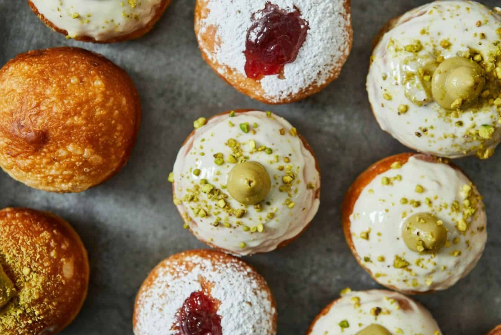 מארז סופגניות אישיות בעבודת יד במילויים אהובים – מגוון סופגניות עגלגלות עם ציפויים כמו אבקת סוכר, פיסטוק קצוץ, זיגוג לבן וריבה, מסודרות על משטח אפור. - Cassarole