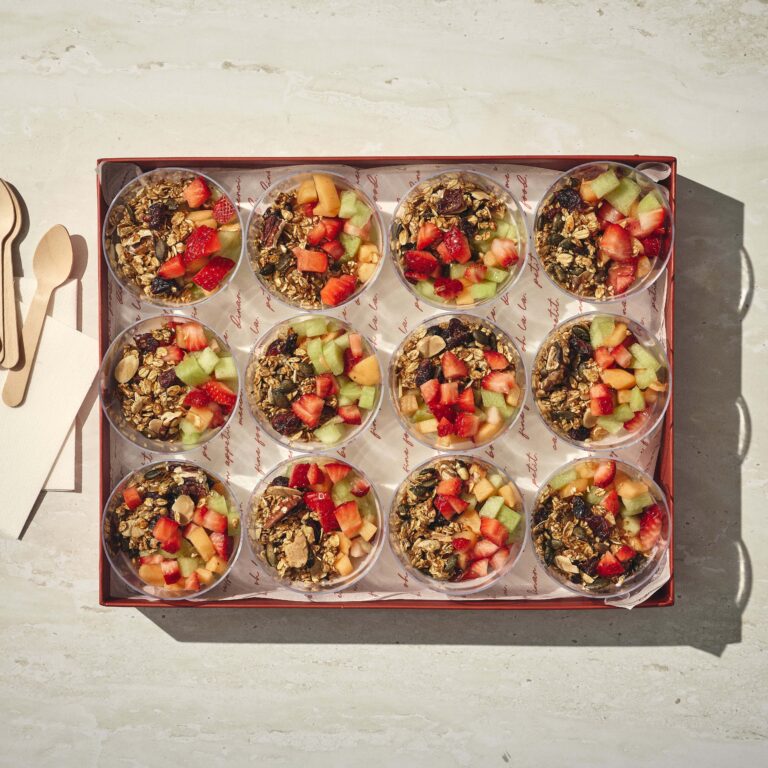 קופסה המכילה 12 כוסות שקופות מלאות בגרנולה, תותים פרוסים וחתכי מלון, עם מפיות וכפיות עץ בצד. - Cassarole