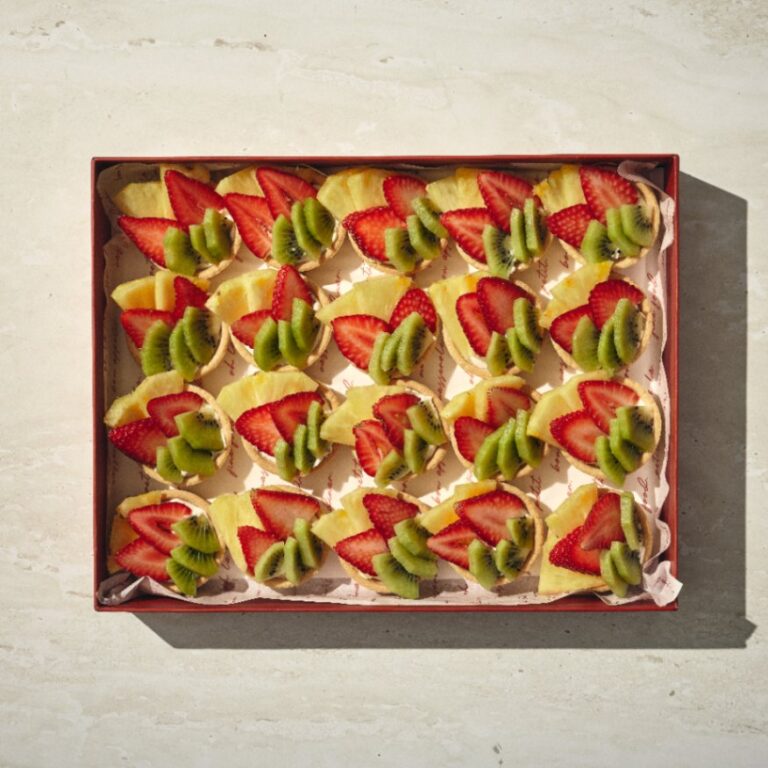 קופסה של טארטלט קיצי — מאפים מרובעים עם פרוסות תותים, קיווי ואננס — מסודרים בשורות מסודרות על משטח בהיר. - Cassarole