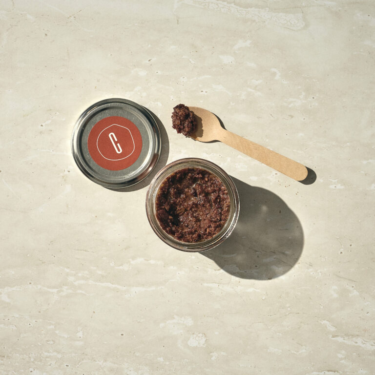 צנצנת זכוכית פתוחה מלאה בממרח כהה ומגושם מונחת ליד המכסה שלה וכף עץ עם מעט ממרח עליה, והכל מונח על משטח אבן בהיר. - Cassarole