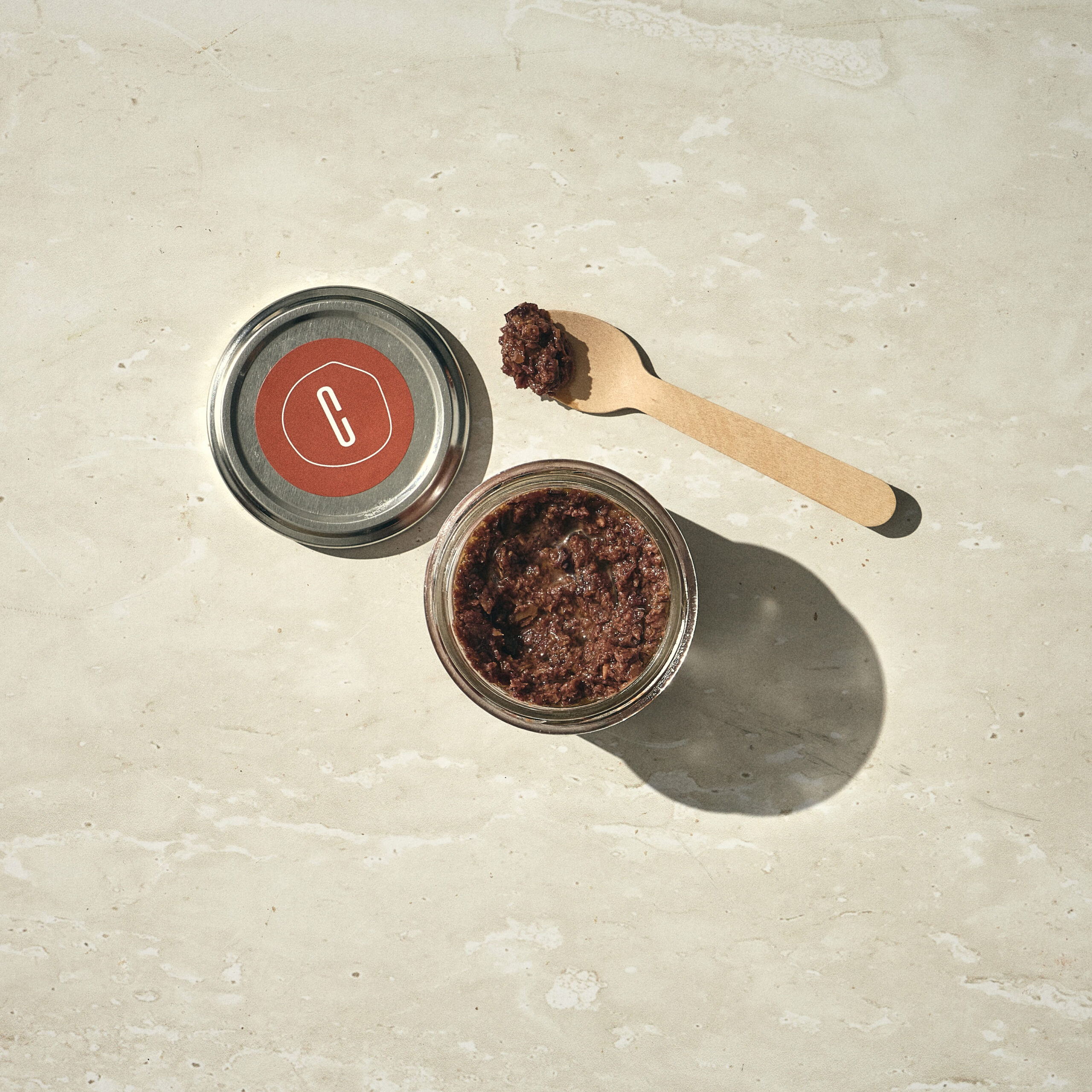 צנצנת זכוכית פתוחה מלאה בממרח כהה ומגושם מונחת ליד המכסה שלה וכף עץ עם מעט ממרח עליה, והכל מונח על משטח אבן בהיר. - Cassarole