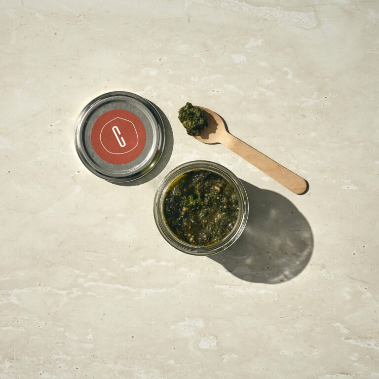 צנצנת זכוכית קטנה מלאה בפסטו ירוק מונחת על משטח אבן בהיר, מכסה המתכת שלה מונח בצד. כפית עץ קטנה עם פסטו מונחת לידה. - Cassarole