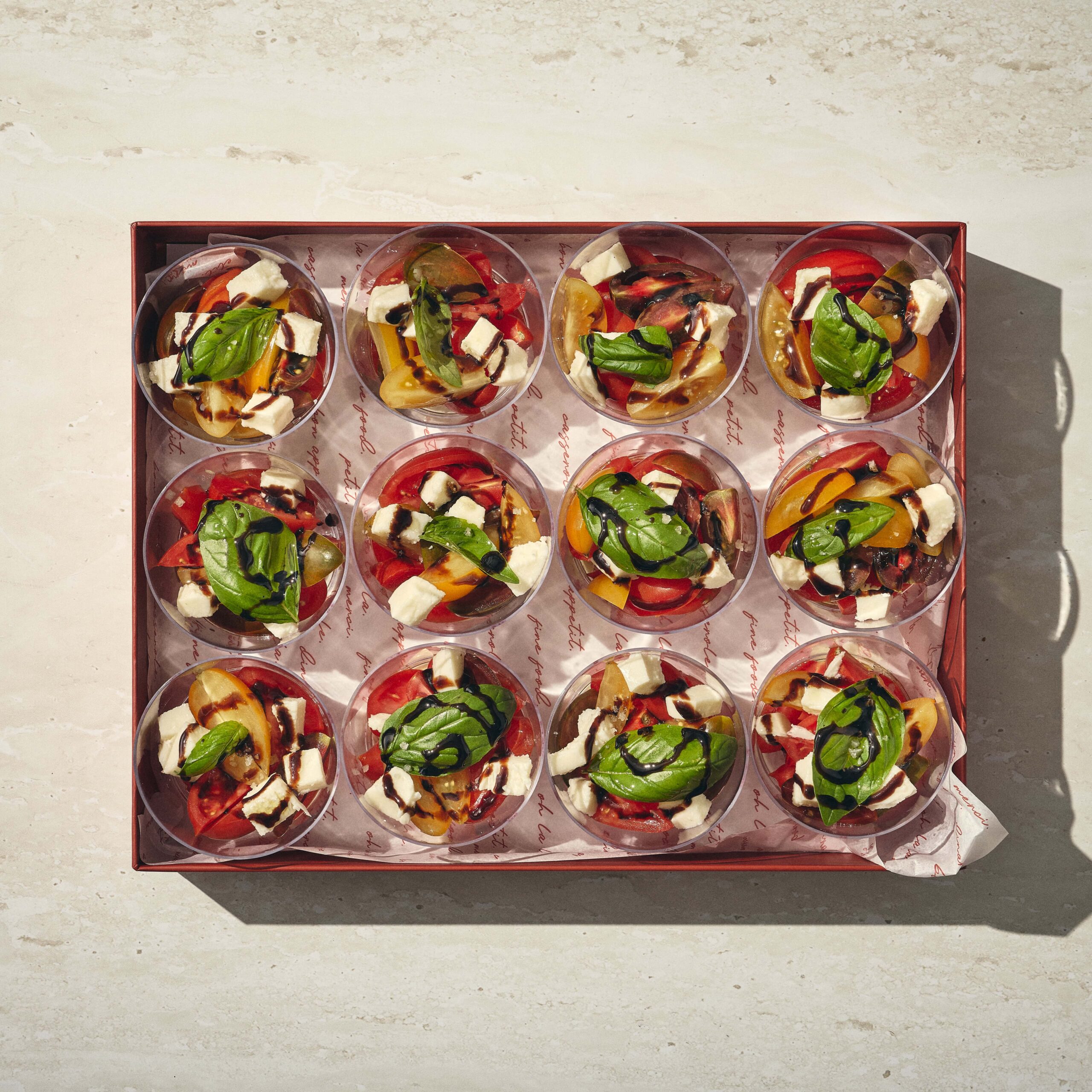 שתים עשרה כוסות מסודרות בקופסה, כל אחת מהן מלאה בסלט עגבניות, מוצרלה ועלי בזיליקום על גבי נייר מעוצב. - Cassarole
