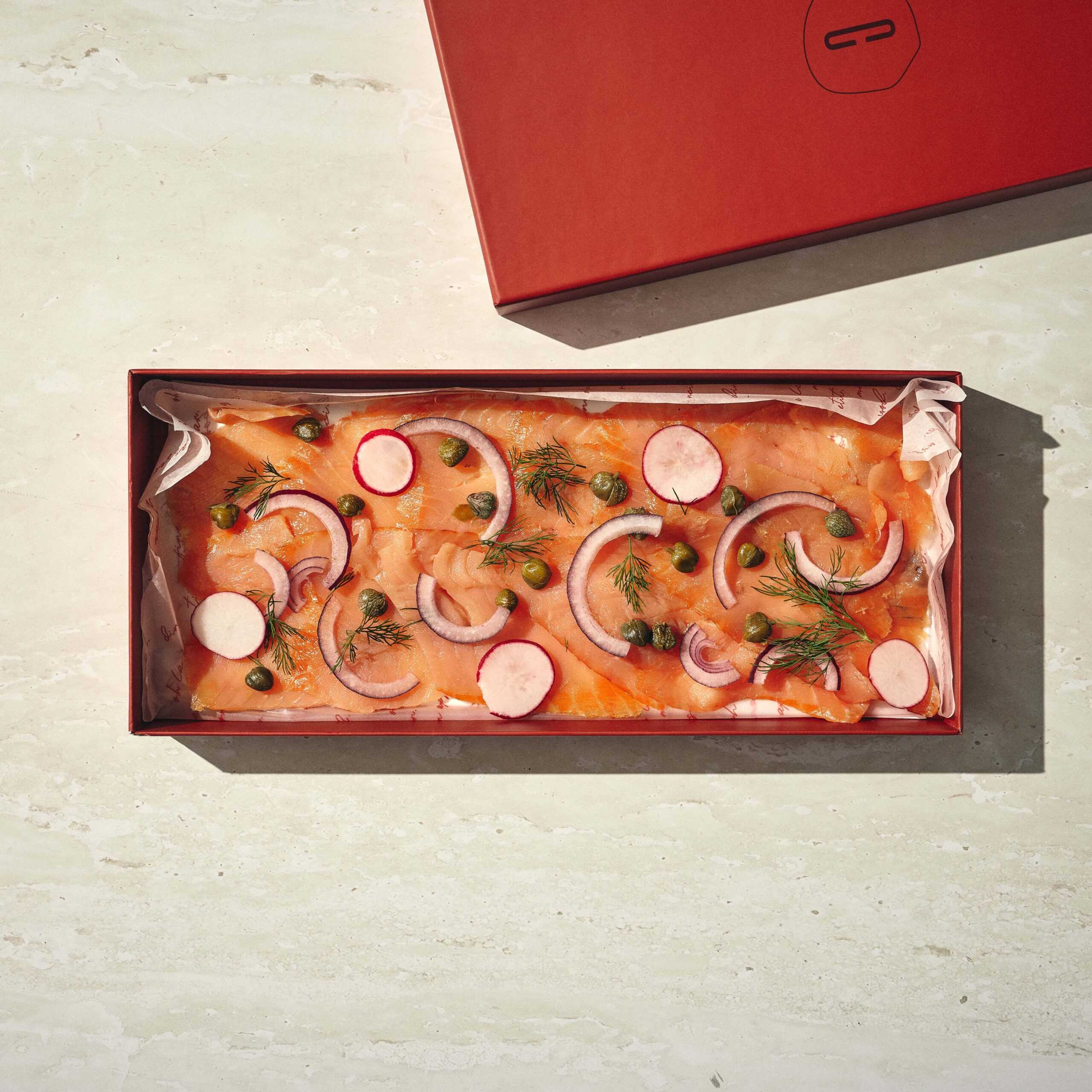 קופסה מלבנית מכילה פרוסות סלמון מעושן עם טבעות בצל סגול, פרוסות צנון, צלפים ושמיר על נייר אפייה, המונחת על משטח בהיר. - Cassarole