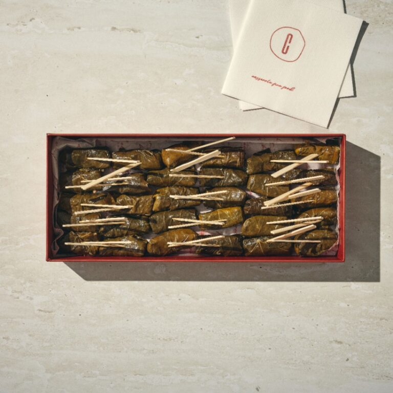 קופסה מלבנית של עלי גפן (גולגולת עלי גפן), כל אחד מהם מהודק בעזרת קיסם עץ, מונחת על משטח בהיר לצד שתי מפיות לבנות מקופלות. - Cassarole