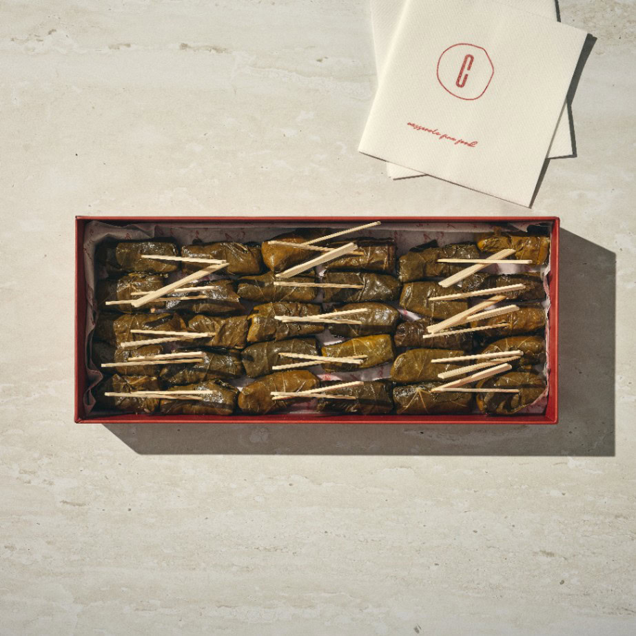 קופסה מלבנית של עלי גפן (גולגולת עלי גפן), כל אחד מהם מהודק בעזרת קיסם עץ, מונחת על משטח בהיר לצד שתי מפיות לבנות מקופלות. - Cassarole