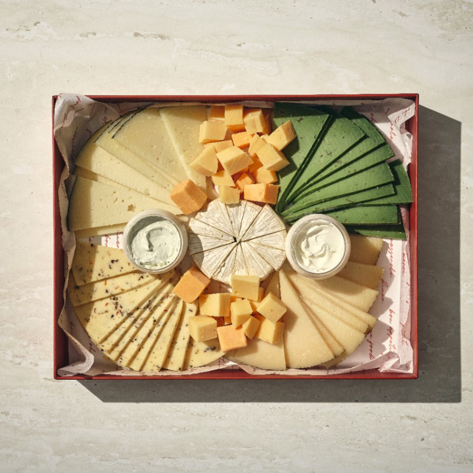 פלטת גבינות בוטיק - גדול: מגש עם גבינות פרוסות וגבינות בקוביות, פרוסות גבינה ירוקה, גלגל גבינה רכה ושני ממרחים קטנים, מסודרים על נייר לבן. - Cassarole