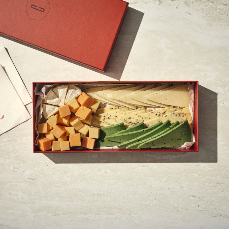פלטת גבינות בוטיק - קטן כוללת קופסה אדומה מלבנית עם פרוסות גבינה במגוון צבעים וצורות - קוביות, משולשים ועיגולים - המסודרות בקפידה על משטח בהיר. - Cassarole