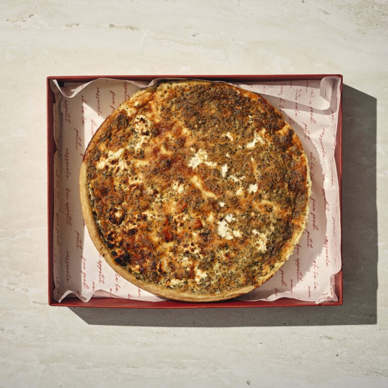 עוגה מלוחה עגולה עם ציפוי חום ומכוסה עשבי תיבול מונחת בקופסה אדומה מרופדת בנייר ממותג על משטח אבן בהיר. - Cassarole