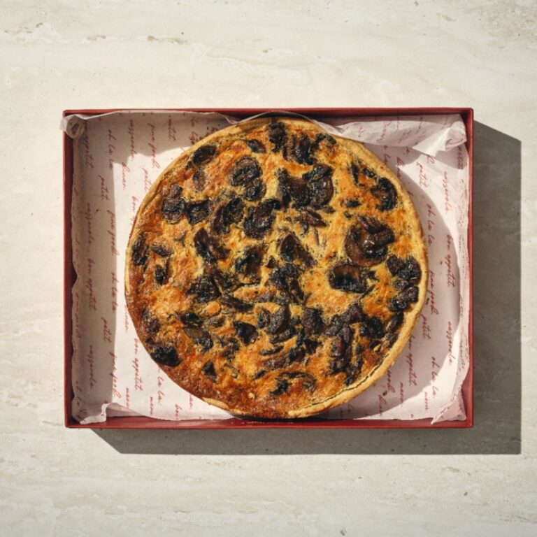 קיש פיטריות עגול מונח בקופסה אדומה פתוחה המרופדת בנייר אפייה מודפס, על גבי משטח בהיר. - Cassarole