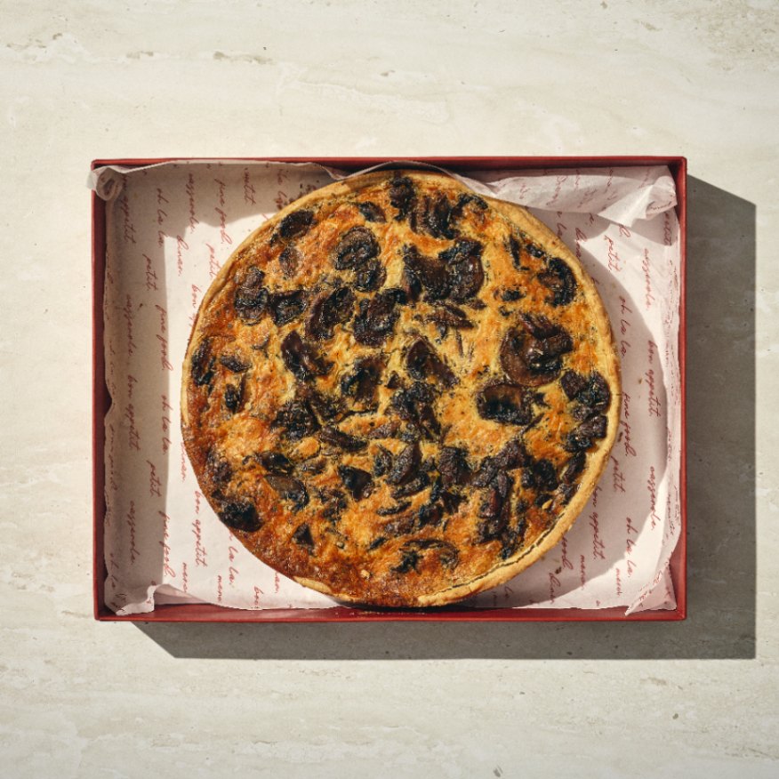 קיש פיטריות עגול מונח בקופסה אדומה פתוחה המרופדת בנייר אפייה מודפס, על גבי משטח בהיר. - Cassarole