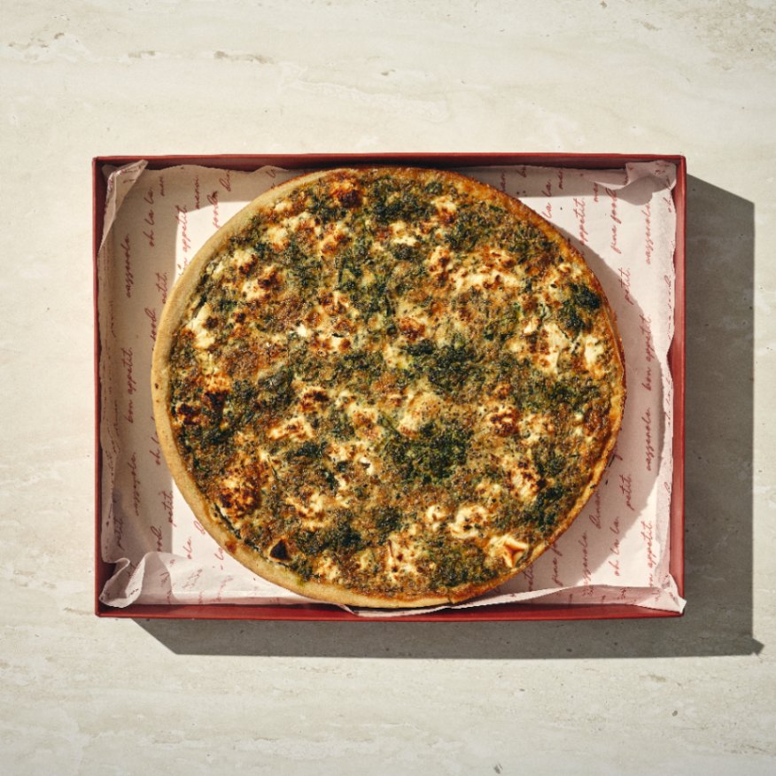 קיש תרד שלם ועגול עם חלק עליון זהוב-חום ועשבי תיבול ירוקים, מוגש בקופסה אדומה פתוחה מרופדת בנייר ממותג על משטח אבן בהיר. - Cassarole