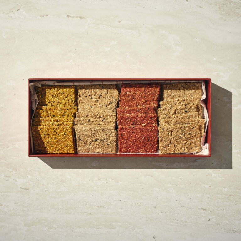 קופסה מלבנית המכילה ארבע שורות של קרקרים מלבניים מסוגים שונים על משטח בהיר. - Cassarole