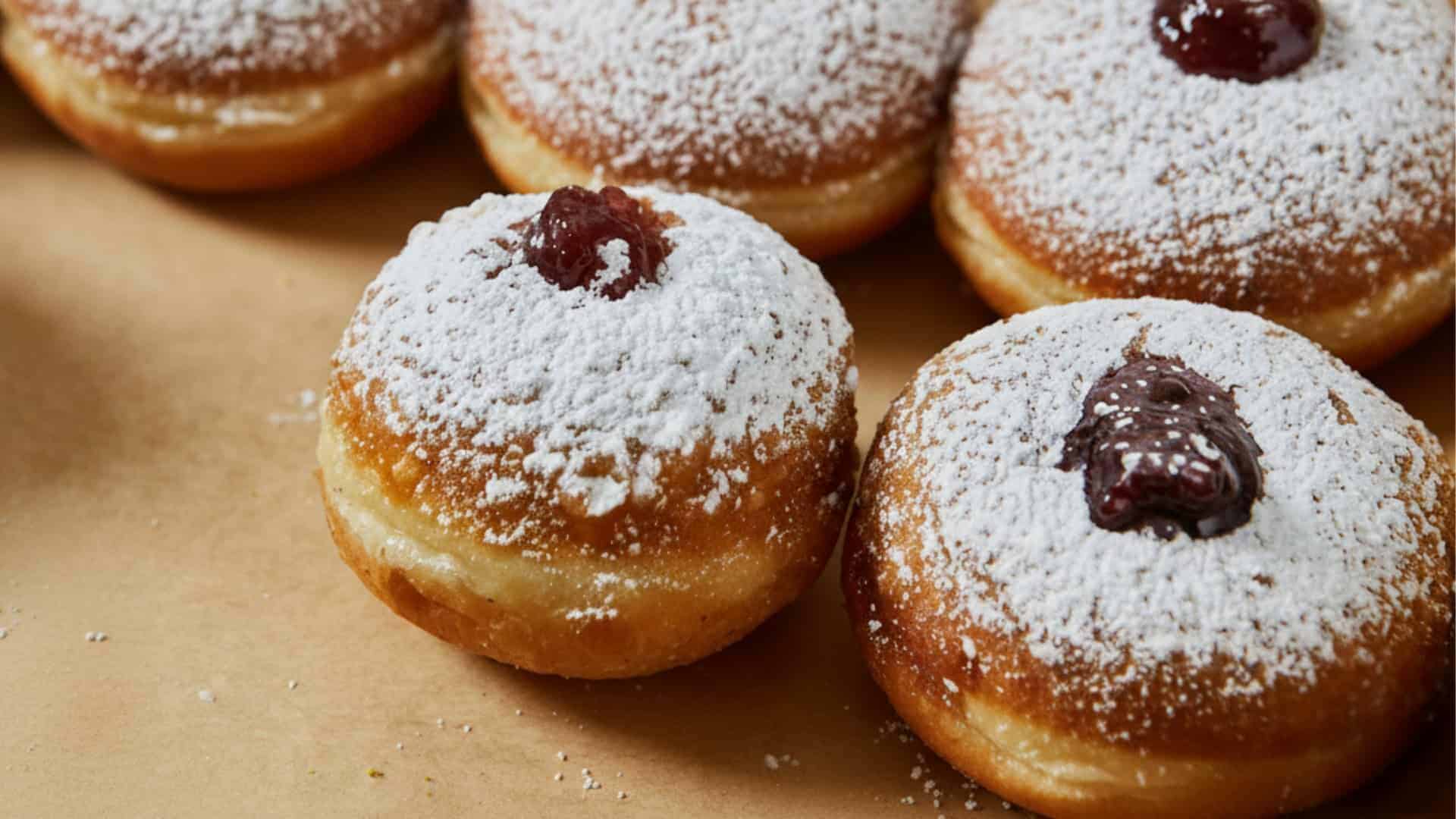 ארבעה סופגניות במילוי ריבה ומצופות אבקת סוכר מסודרות על משטח חום. - Cassarole